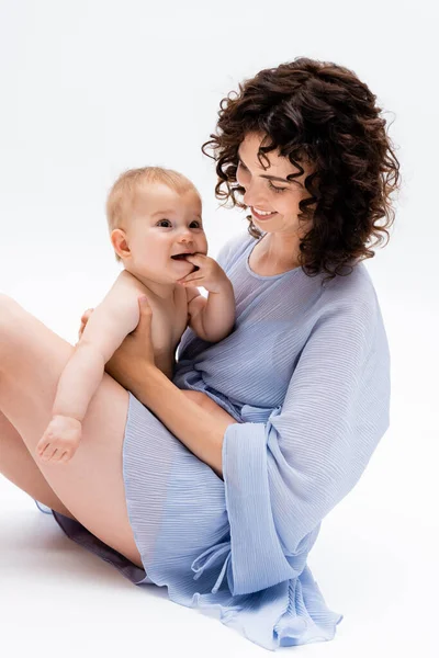 Sorrindo mãe no vestido olhando para a filha bebê satisfeito enquanto sentado no fundo branco — Fotografia de Stock