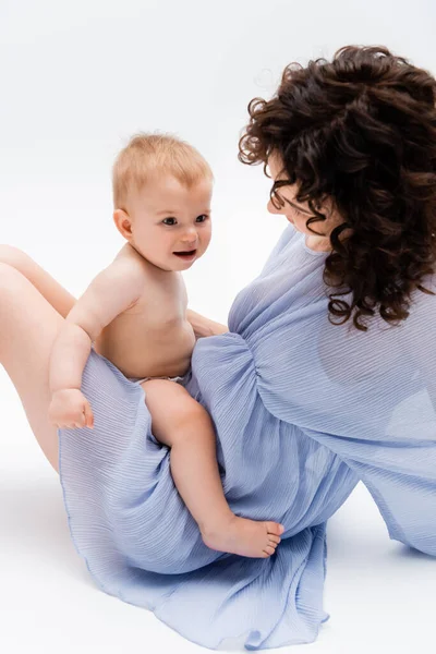 Lockige Mutter im blauen Kleid schaut fröhliches Baby-Mädchen auf weißem Hintergrund an — Stockfoto