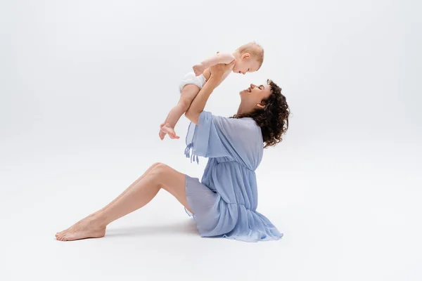 Vue latérale de heureux pieds nus maman en robe jouer avec bébé fille sur fond blanc — Photo de stock