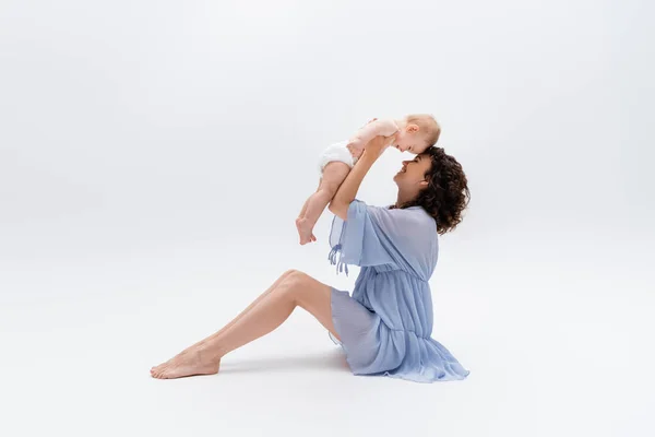 Vista lateral de mamá alegre en vestido sosteniendo hija bebé sobre fondo blanco - foto de stock