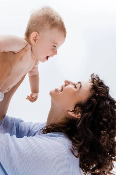 Brünette Mutter in blauem Kleid schaut fröhliche Säuglingstochter isoliert auf weißem Grund an — Stockfoto