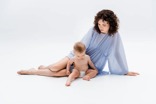 Barefoot woman in dress looking at baby girl sitting on white background — Stock Photo