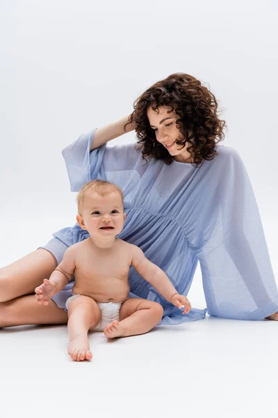 Alegre niña sentada cerca de la mamá rizada en vestido sobre fondo blanco - foto de stock