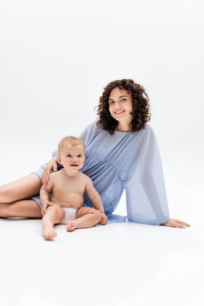 Glückliche Frau in blauem Kleid blickt in die Kamera neben einem Säugling, der auf weißem Hintergrund sitzt — Stockfoto