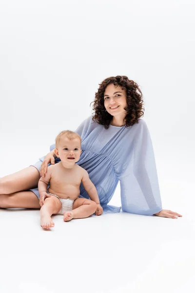 Donna felice in abito blu guardando la fotocamera vicino bambino seduto su sfondo bianco — Foto stock