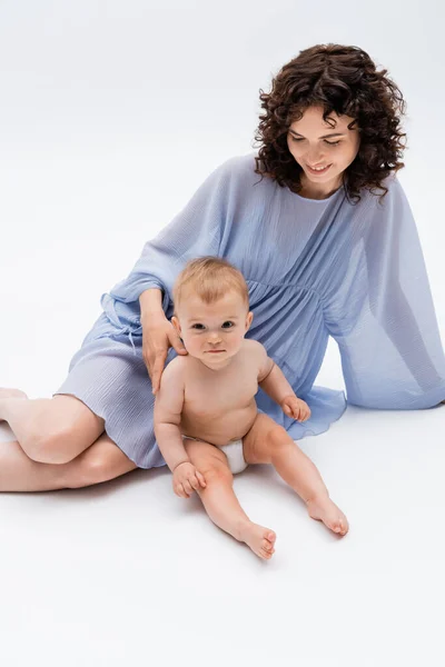 Positive Mutter im Kleid sitzt in der Nähe der kleinen Tochter und schaut in die Kamera auf weißem Hintergrund — Stockfoto