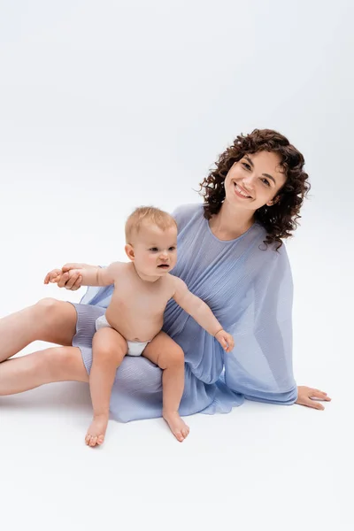 Madre en vestido azul cogida de la mano de la hija lactante y mirando a la cámara sobre fondo blanco - foto de stock