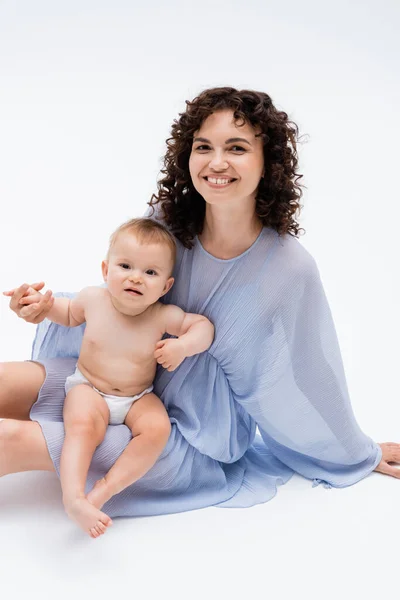 Happy woman holding hand of baby daughter and sitting on white background — Stock Photo