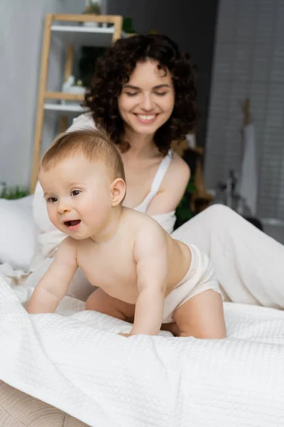 Baby daughter crawling on bed near blurred mother at home — Stock Photo