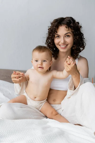 Happy curly mother holding hands of infant daughter on bed — Stock Photo