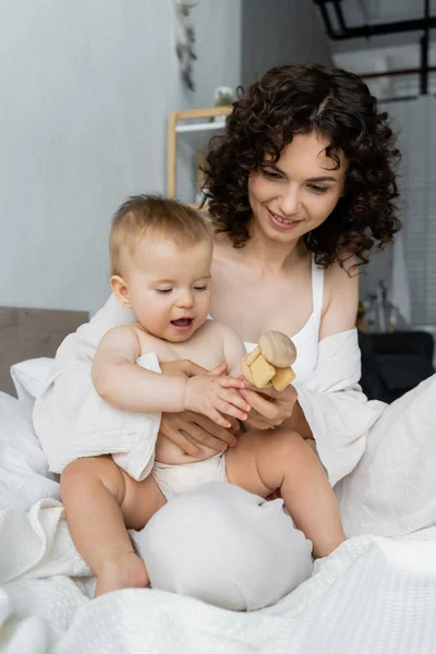 Curly mother holding toy near infant daughter on bed — Stock Photo