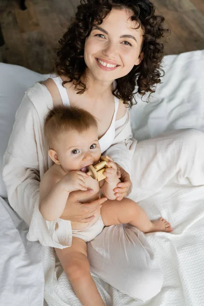 High angle view of curly parent looking at camera near baby daughter with wooden toy on bed — Stock Photo