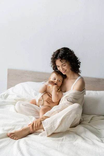 Улыбающаяся женщина обнимает дочку с деревянной игрушкой на кровати — стоковое фото