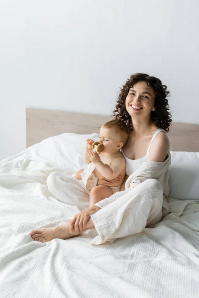 Позитивная брюнетка родитель обнимает ребенка с деревянной игрушкой на кровати — стоковое фото
