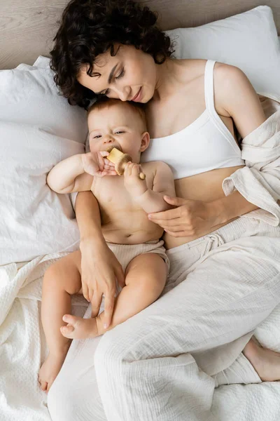 Visão superior da mulher encaracolada em pijama tocando bebê menina segurando brinquedo de madeira na cama — Fotografia de Stock