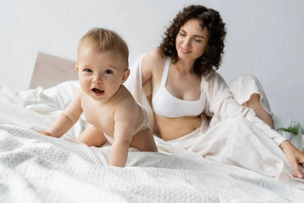 Joyeux bébé fille regardant la caméra près du parent flou en pyjama sur le lit — Photo de stock