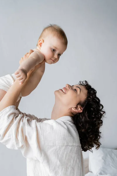 Curly woman smiling while lifting baby girl in bedroom in morning — Stock Photo