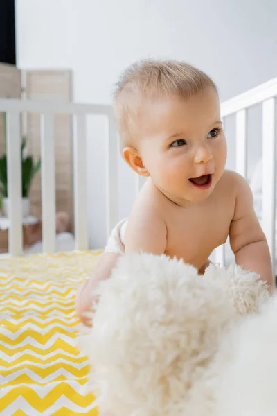 Exited baby girl looking away near soft toy in crib — Stock Photo