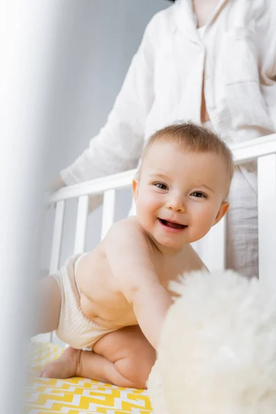 Bébé fille heureuse en culotte regardant la caméra près floue jouet mou dans la crèche et la mère à la maison — Photo de stock