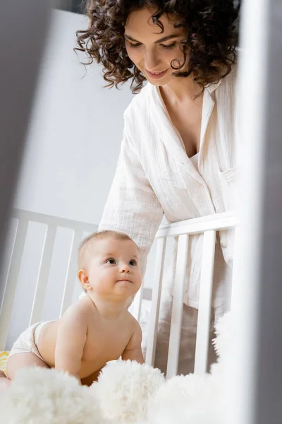 Femme souriante regardant bébé fille près flou jouet mou dans la crèche — Photo de stock