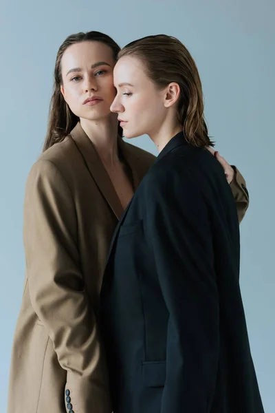 Young woman in beige blazer hugging lesbian partner and looking at camera isolated on grey — Stock Photo