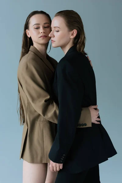 Brünette Frau im beigen Blazer umarmt junge lesbische Freundin isoliert auf grau — Stockfoto