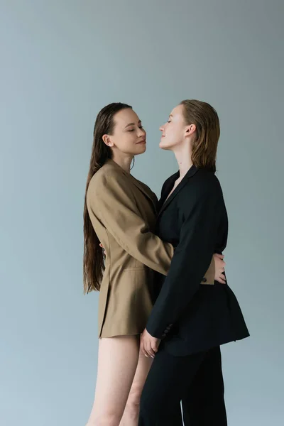 Hübsche Frau mit langen Haaren umarmt lesbische Freundin in schwarzem Blazer isoliert auf grau — Stockfoto