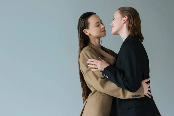 Young and happy lesbian women in blazers embracing face to face isolated on grey — Stock Photo