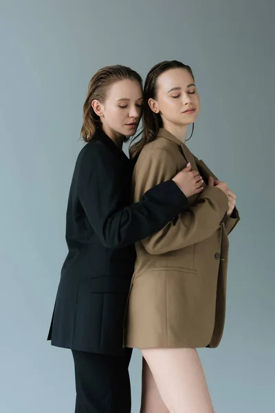 Giovane e tenera donna che abbraccia la ragazza lesbica in piedi con gli occhi chiusi isolati sul grigio — Foto stock