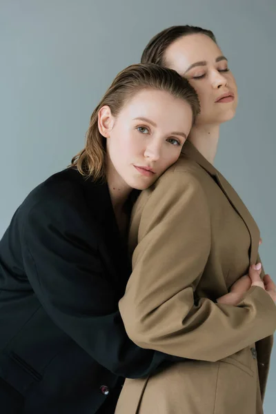 Pretty lesbian woman looking at camera and embracing lesbian girlfriend standing with closed eyes isolated on grey — Stock Photo