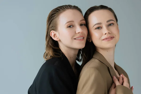 Jeune et heureux couple de même sexe en beige et noir vestes souriant à la caméra isolé sur gris — Photo de stock