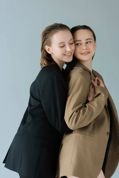 Веселая лесбиянка обнимает счастливую молодую девушку в бежевом пиджаке, изолированном от серых — стоковое фото
