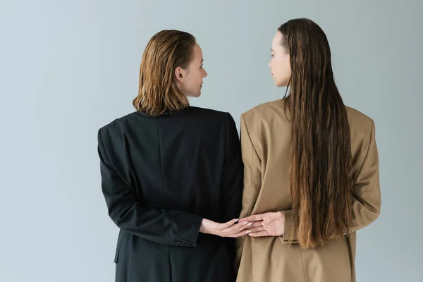 Vista trasera de mujeres lesbianas en chaquetas negras y beige mirándose y tomándose de las manos aisladas en gris - foto de stock