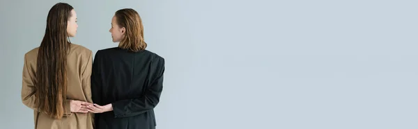 Back view of lesbian women in blazers holding hands and looking at each other isolated on grey, banner — Stock Photo