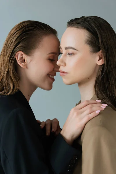 Side view of young woman touching shoulder of lesbian girlfriend and smiling isolated on grey — Stock Photo