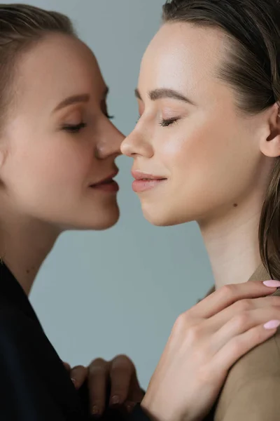 Vista lateral de las amigas lesbianas sonrientes de pie cara a cara con los ojos cerrados aislados en gris - foto de stock