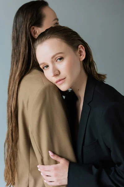 Young lesbian woman looking at camera while leaning on long haired lesbian girlfriend isolated on grey — Stock Photo