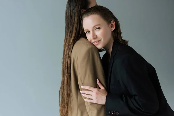 Mujer lesbiana complacida sonriendo a la cámara y abrazando a su novia con el pelo largo aislado en gris - foto de stock
