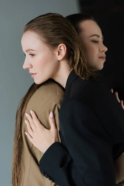 Vista lateral de mujeres lesbianas jóvenes y bonitas en blazers abrazando sobre fondo gris - foto de stock