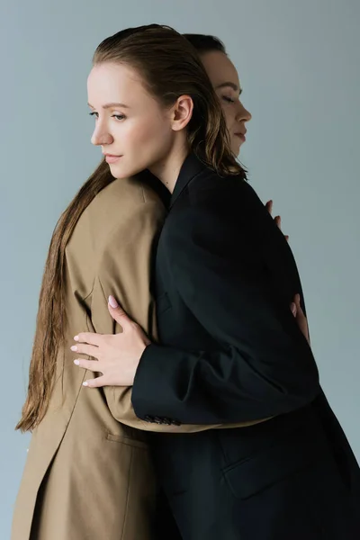Young and pretty same sex couple in blazers hugging isolated on grey — Stock Photo