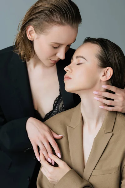 Sensual lésbica mulher com olhos fechados abraçando parceiro no bege blazer isolado no cinza — Fotografia de Stock