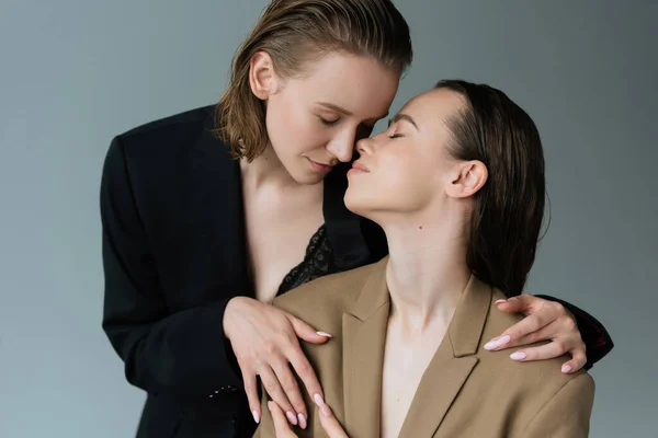 Sinnliche Frau im schwarzen Blazer umarmt brünette lesbische Freundin isoliert auf grau — Stockfoto