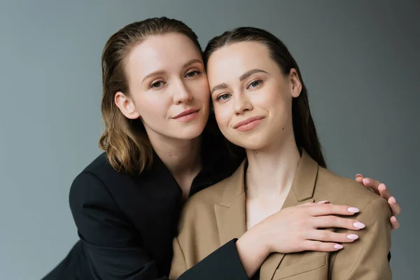 Young and pleased lesbian partners in beige and black jackets looking at camera isolated on grey — Stock Photo