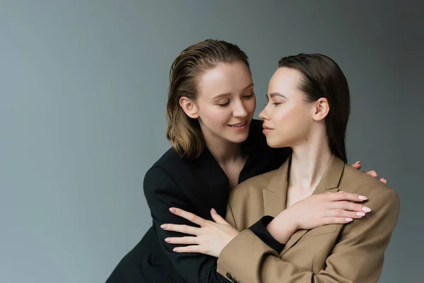 Lesbiche sorridenti in blazer beige e neri che si abbracciano isolate sul grigio — Foto stock