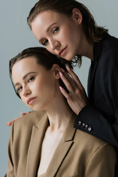 Young and sensual lesbian couple in blazers looking at camera isolated on grey — Stock Photo