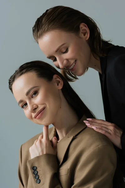 Bonita mujer morena en blazer beige mirando a la cámara cerca de feliz novia lesbiana aislada en gris - foto de stock