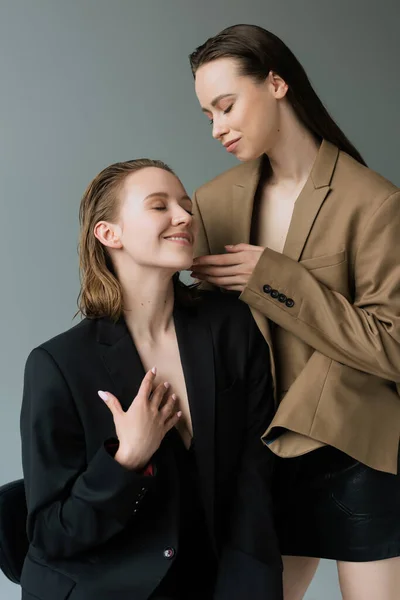 Brunette woman touching face of happy girlfriend sitting with closed eyes isolated on grey — Stock Photo