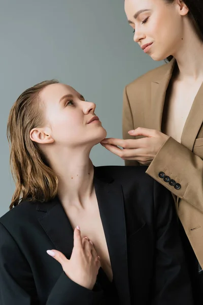 Jeune femme en blazer beige touchant menton de jeune copine lesbienne isolé sur gris — Photo de stock