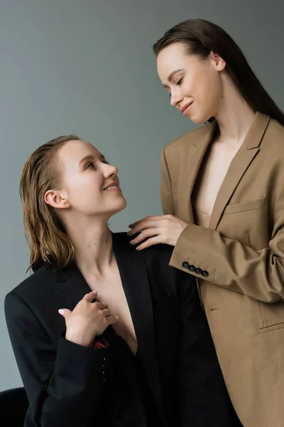 Young and pretty lesbian women in beige and black blazers smiling at each other isolated on grey — Stock Photo