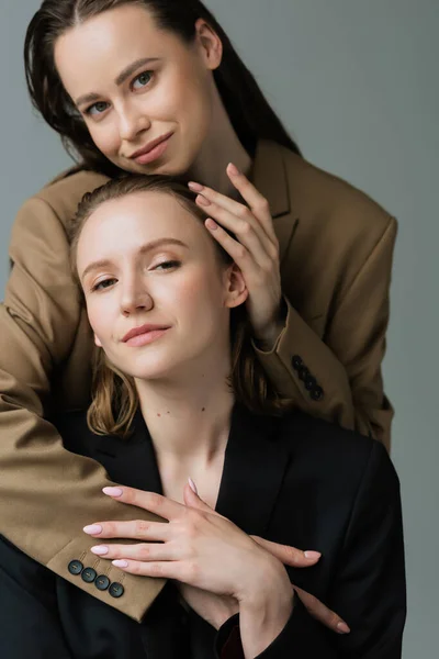 Young and pleased lesbian partners in blazers looking at camera and embracing isolated on grey — Stock Photo